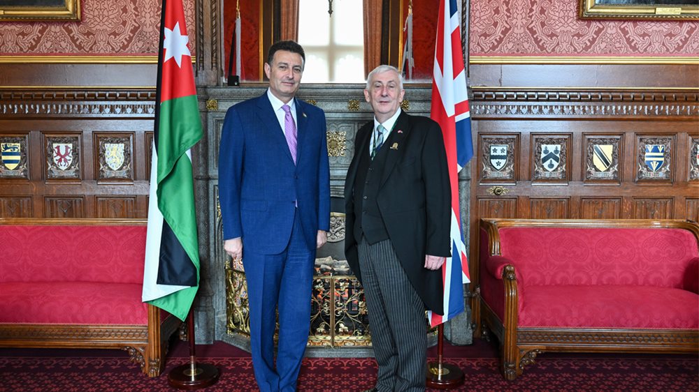 The Speaker of the House of Commons and the  Speaker of Jordan standing side by side in front of an ornate fireplace. The flags of Jordan and the United  Kingdom are at their sides.