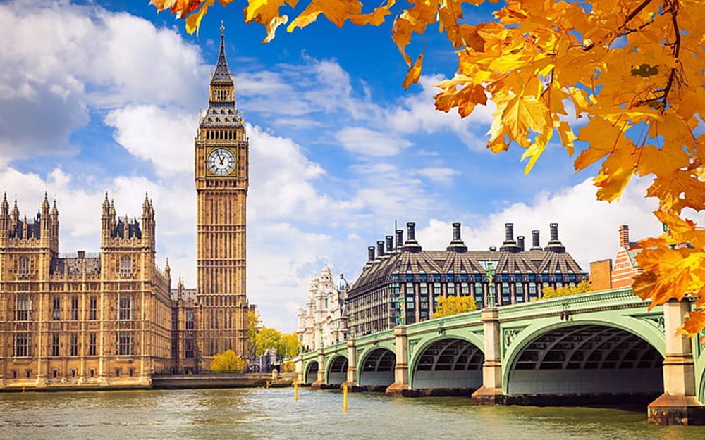 Image looking across the River Thames in London at Westminster Bridge towards the iconic Big Ben, the Palace of Westminster and Portcullis House, framed with vibrant autumn foliage and a bright blue sky with clouds