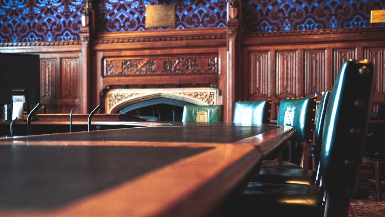A wood panelled room with an ornate fireplace. A large wooden table with a black top is in the foreground. Green leather upright chairs are to the right of the table.
