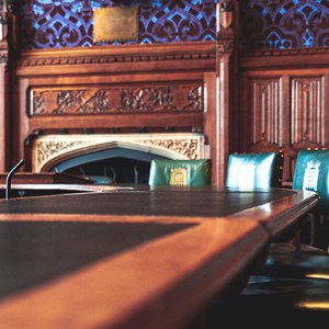 A wood panelled room with an ornate fireplace. A large wooden table with a black top is in the foreground. Green leather upright chairs are to the right of the table.
