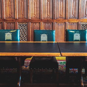 A wood panelled room with five green leather chairs behind a wooden table. There is a gold portcullis on the back of the chairs.