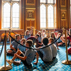 A group of school children wearing headsets, sitting on a blue carpet in an ornately decorated room A woman in a red dress is kneeling with the group and is explaining something to the children.