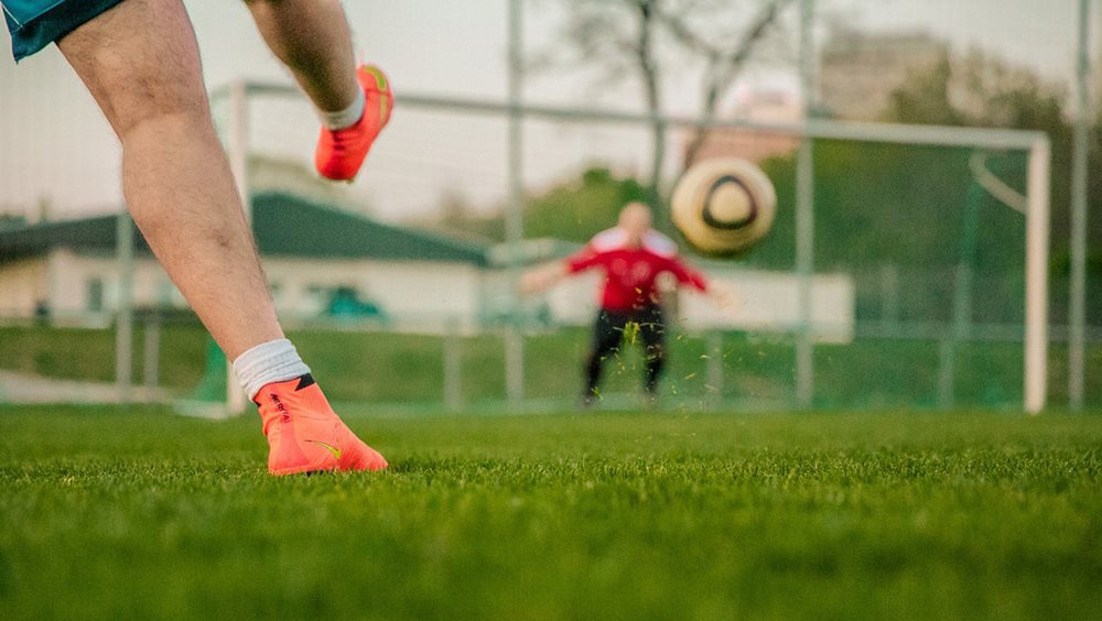 A football shot towards a goal. A man's legs are on the left of the image , he is wearing bright orange boots. The goalkeeper is wearing red and the ball is in front of the goal in mid air.