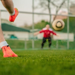 A football shot towards a goal. A man's legs are on the left of the image , he is wearing bright orange boots. The goalkeeper is wearing red and the ball is in front of the goal in mid air.
