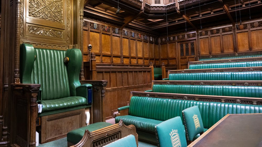 The Speaker's chair, a large green leather chair with a carved wooden canopy, is on the left of the image. Green House of Commons benches are on the right and carved wooden panelling is in the background.